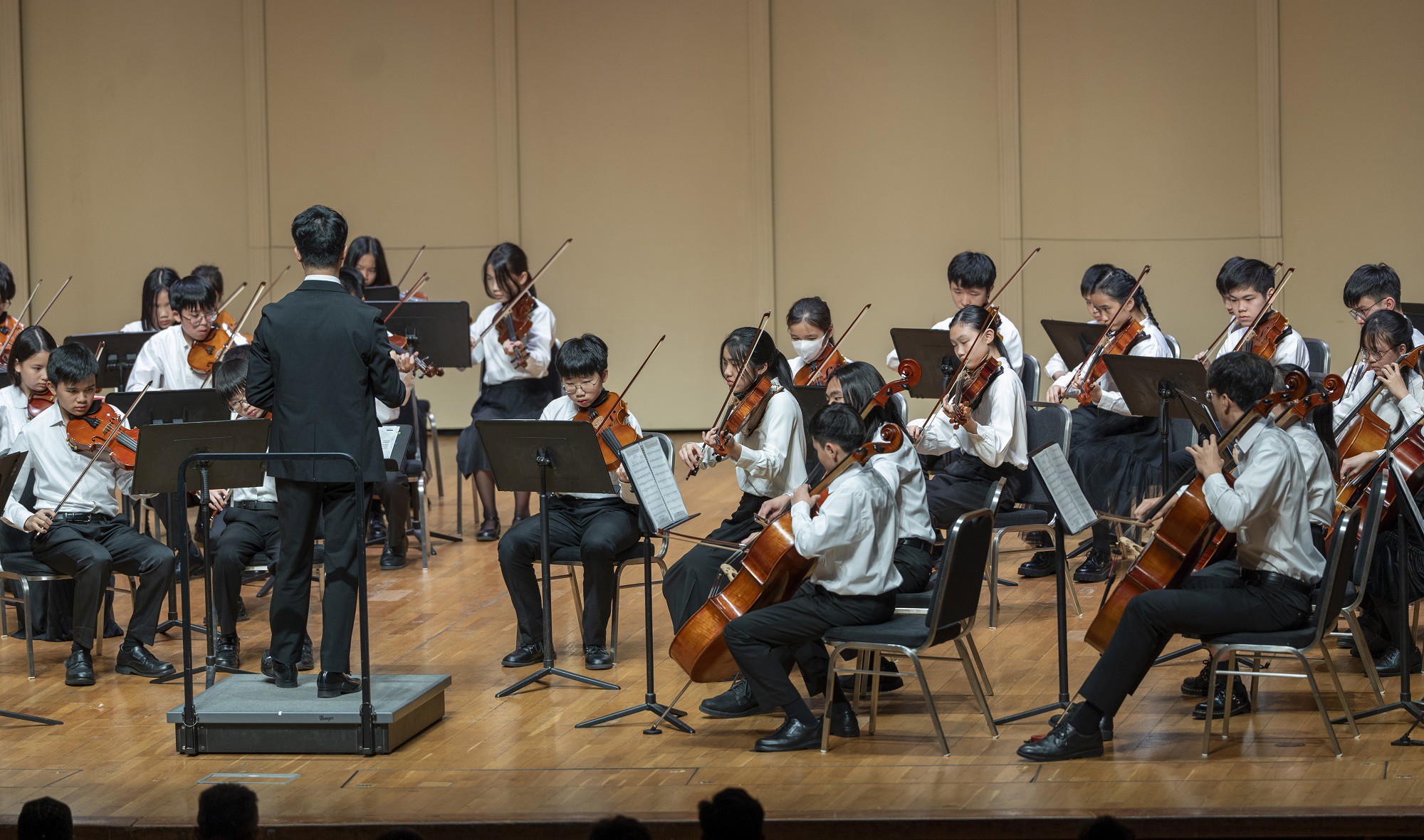 學生管弦樂團在音樂廳演出，指揮帶領年輕樂手演奏弦樂器。